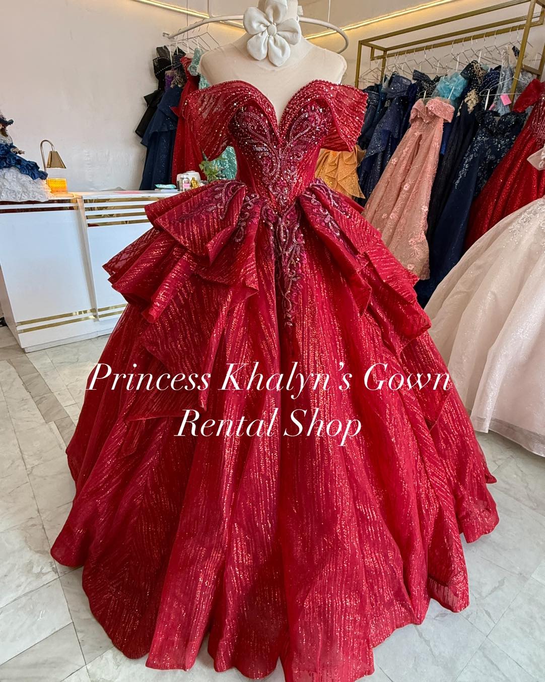 A stunning red off-shoulder ball gown with intricate beadwork and shimmering fabric. The gown features a fitted bodice with elaborate embroidery and a voluminous skirt adorned with layered ruffles, creating a regal and dramatic silhouette.