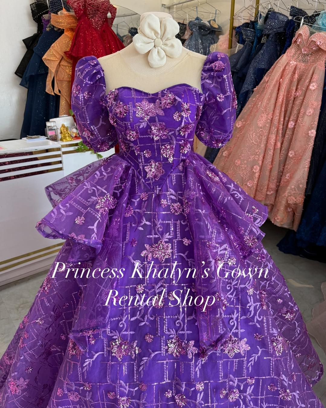 A vibrant purple ball gown with puffed sleeves and intricate pink floral embroidery. The gown features a sweetheart neckline and a voluminous layered skirt, creating a dramatic and elegant silhouette.
