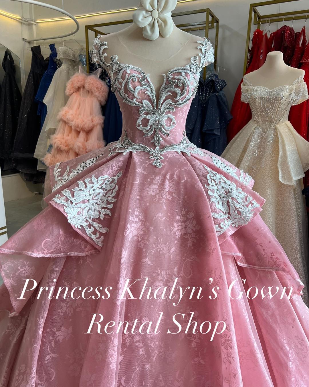 A stunning pink ball gown with an intricate floral lace bodice and illusion neckline. The gown features elaborate white embroidery and a voluminous layered skirt, perfect for formal occasions.