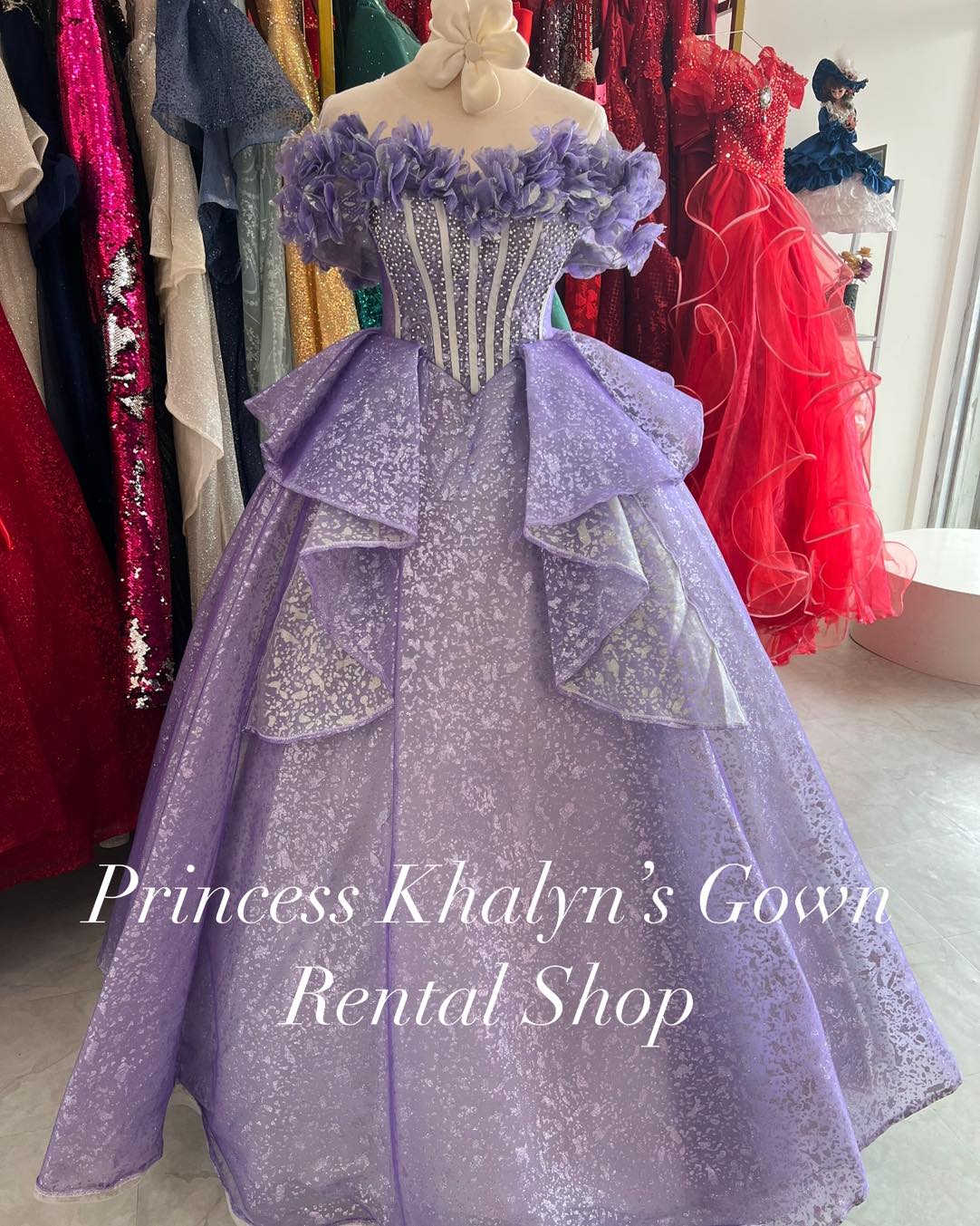 A stunning lavender ball gown with off-shoulder floral appliqué on the bodice, a structured corset with bead embellishments, and a voluminous shimmering skirt with layered ruffles.