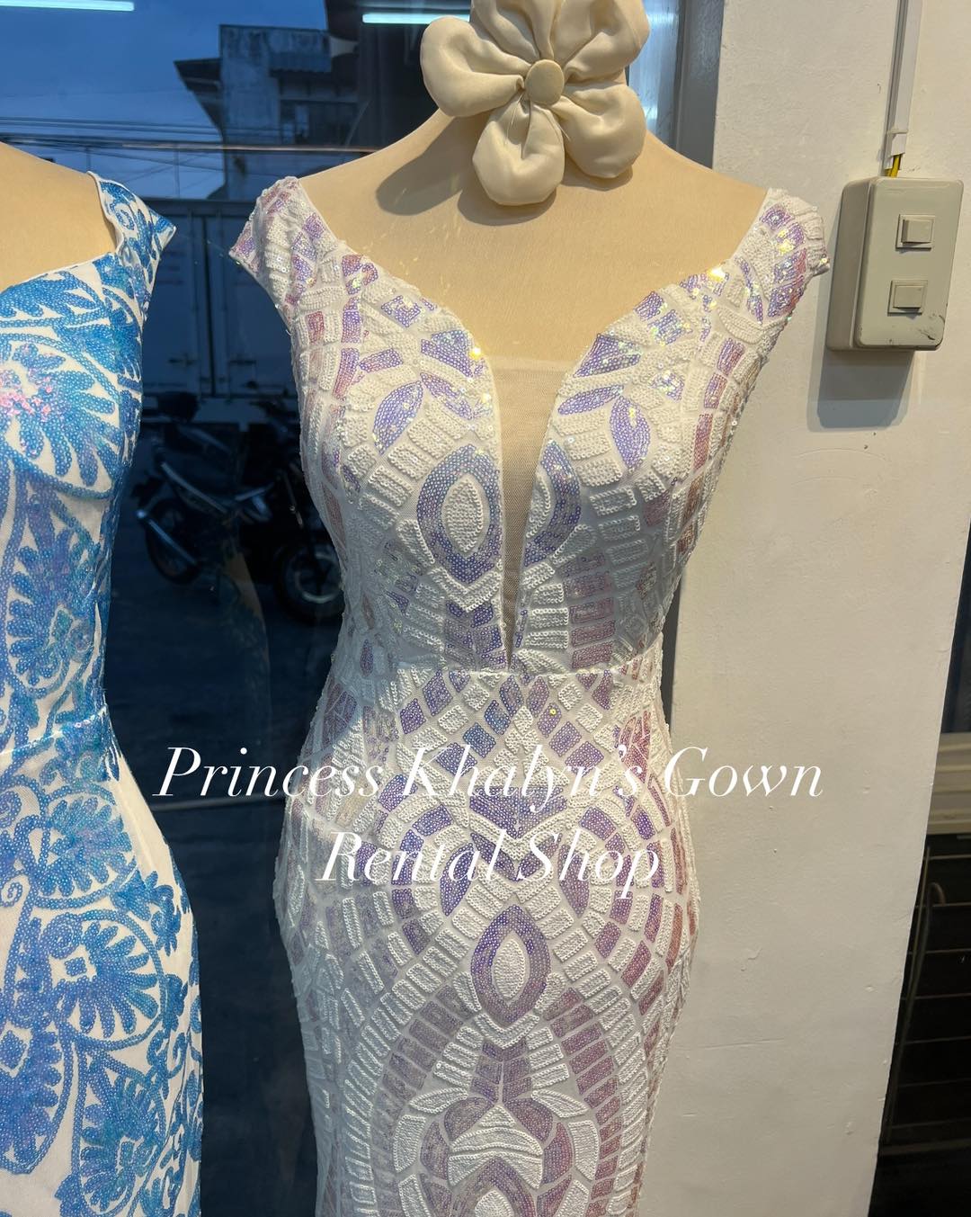 A sophisticated gown featuring a deep V-neckline with cap sleeves. The dress is adorned with intricate lavender and white lace patterns, creating an elegant and textured appearance. Suitable for formal events and special occasions.