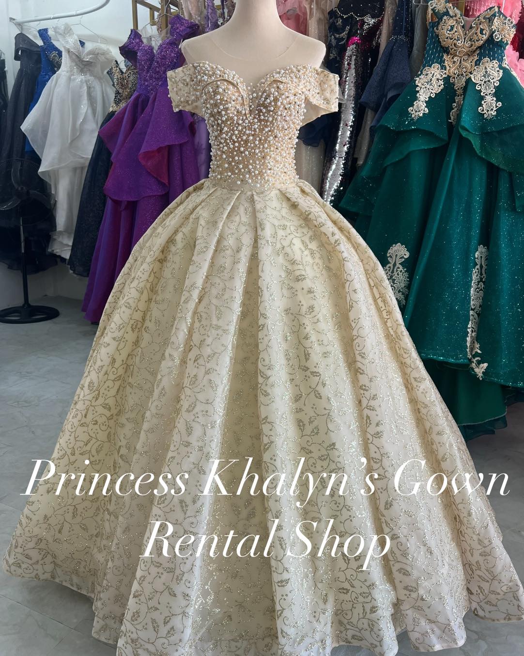 A luxurious gold off-shoulder ball gown adorned with intricate pearl beadwork on the bodice and a voluminous skirt featuring delicate swirling embroidery. The gown exudes a regal and sophisticated charm, suitable for formal occasions.