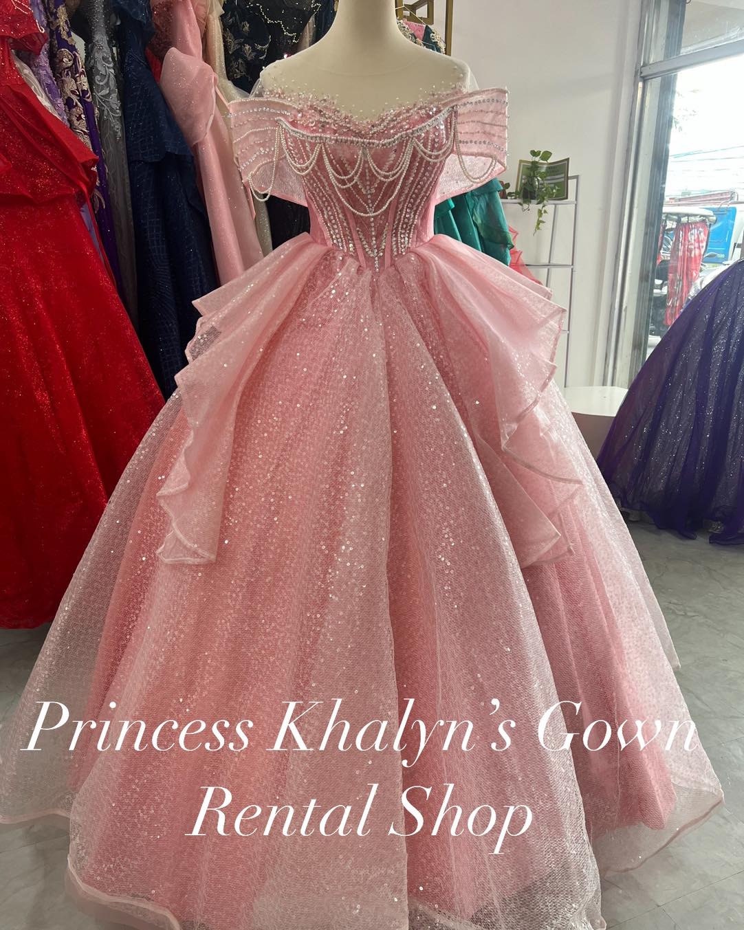 A pink off-shoulder ball gown with a shimmering textured fabric and intricate pearl beadwork cascading across the bodice. The gown features a voluminous skirt with layered ruffles, creating a dreamy and elegant silhouette.