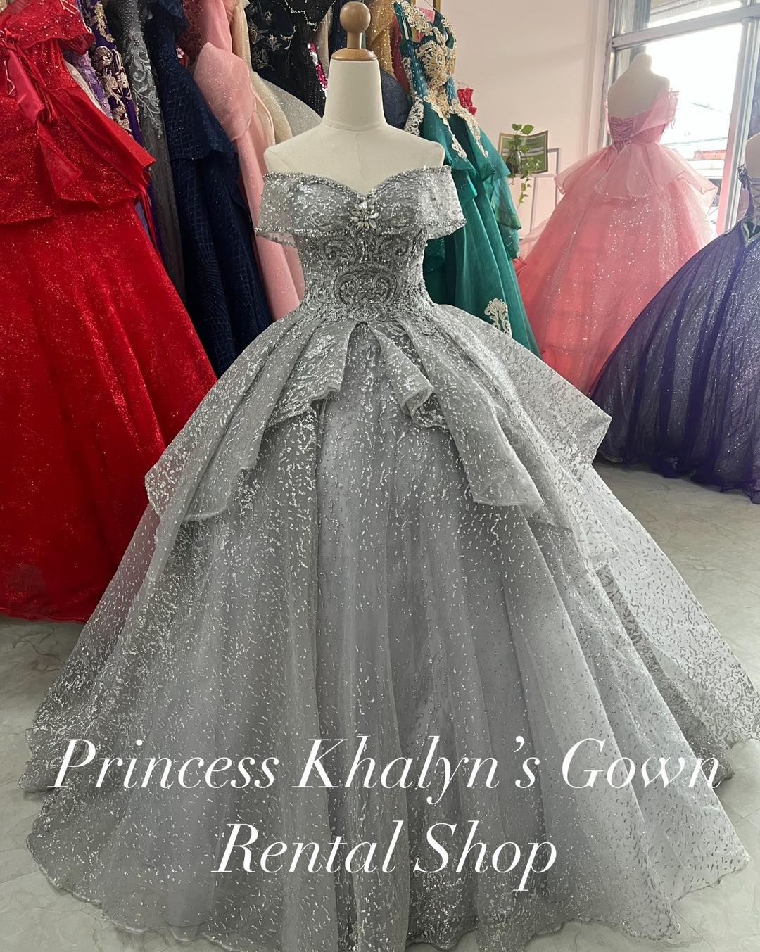 A dazzling silver off-shoulder ball gown with intricate beadwork and a voluminous layered skirt. The gown features a fitted bodice adorned with shimmering details, creating an elegant and sophisticated look.