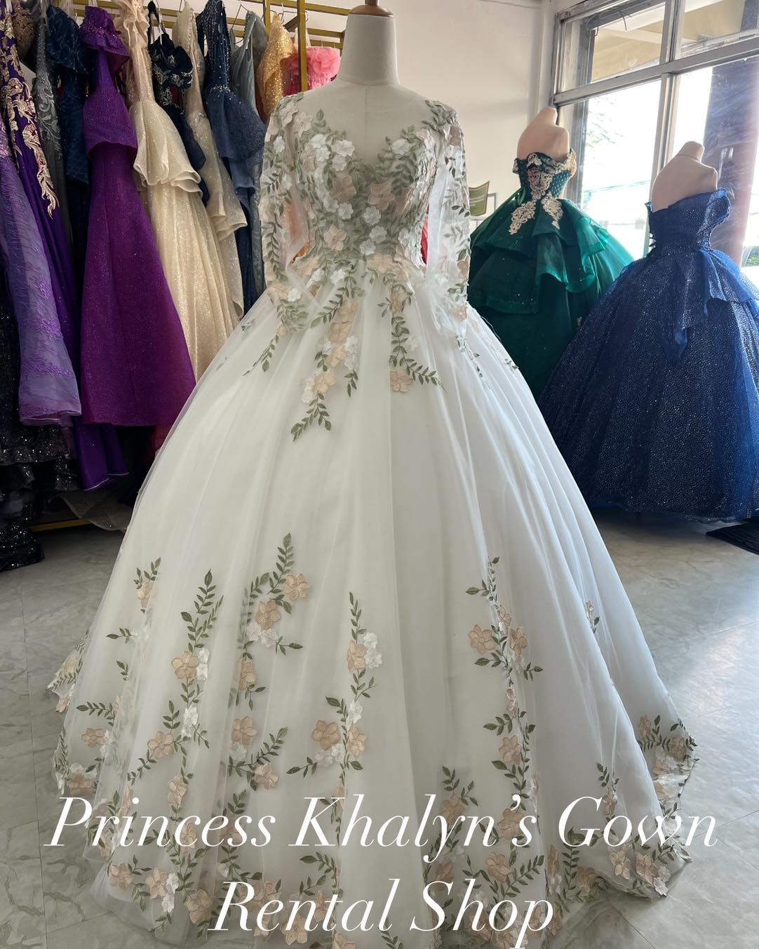 A white ball gown with long sheer sleeves and intricate floral appliqué in soft pastel colors. The gown features a fitted bodice with delicate embroidery and a voluminous skirt adorned with cascading floral patterns, creating an elegant and romantic look.