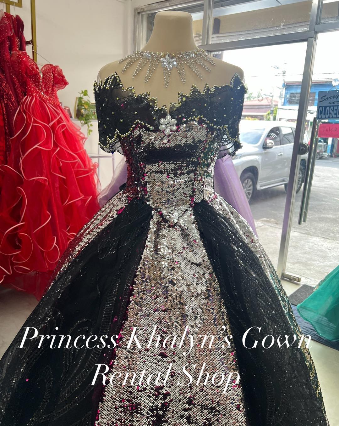 A dazzling black off-shoulder gown adorned with shimmering silver sequins and intricate floral embellishments. The gown features a dramatic neckline with beaded details and a voluminous skirt, perfect for making a statement at any formal event.