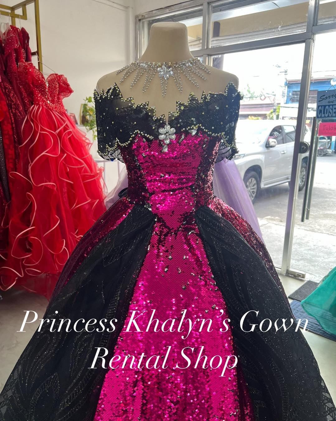 A striking fuchsia pink gown with shimmering sequins and black lace detailing. The gown features an off-shoulder design with intricate beadwork and floral embellishments on the bodice, complemented by a voluminous black overlay skirt.