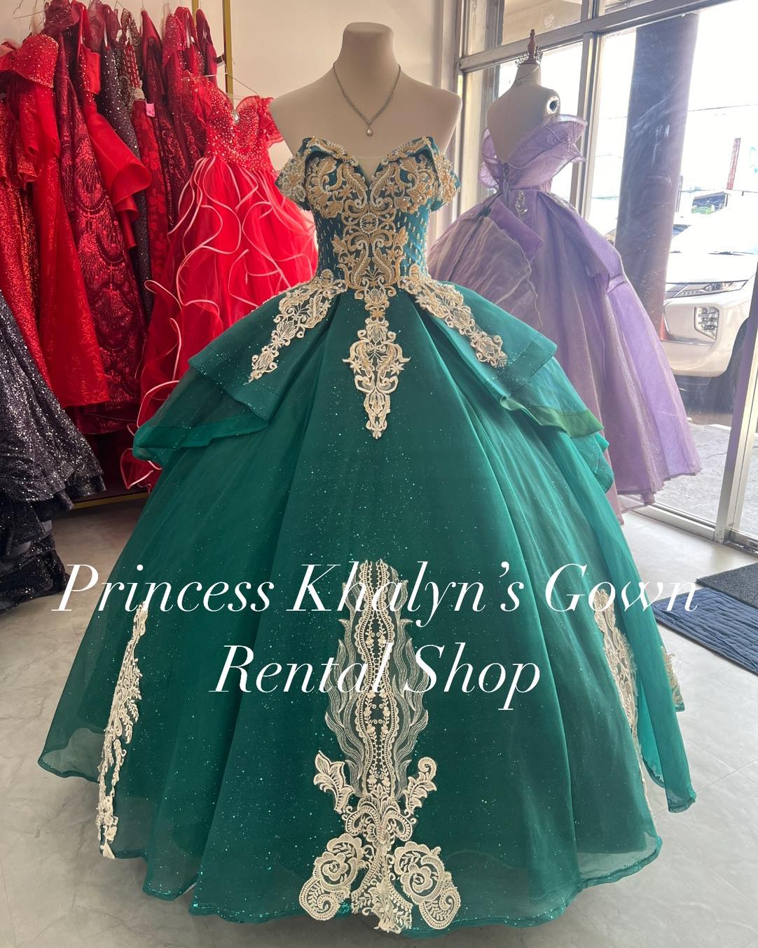 A stunning emerald green ball gown with an off-shoulder design, featuring intricate gold embroidery on the bodice and cascading down the voluminous skirt. The gown is adorned with shimmering details and layered tulle, creating a regal and elegant appearance.