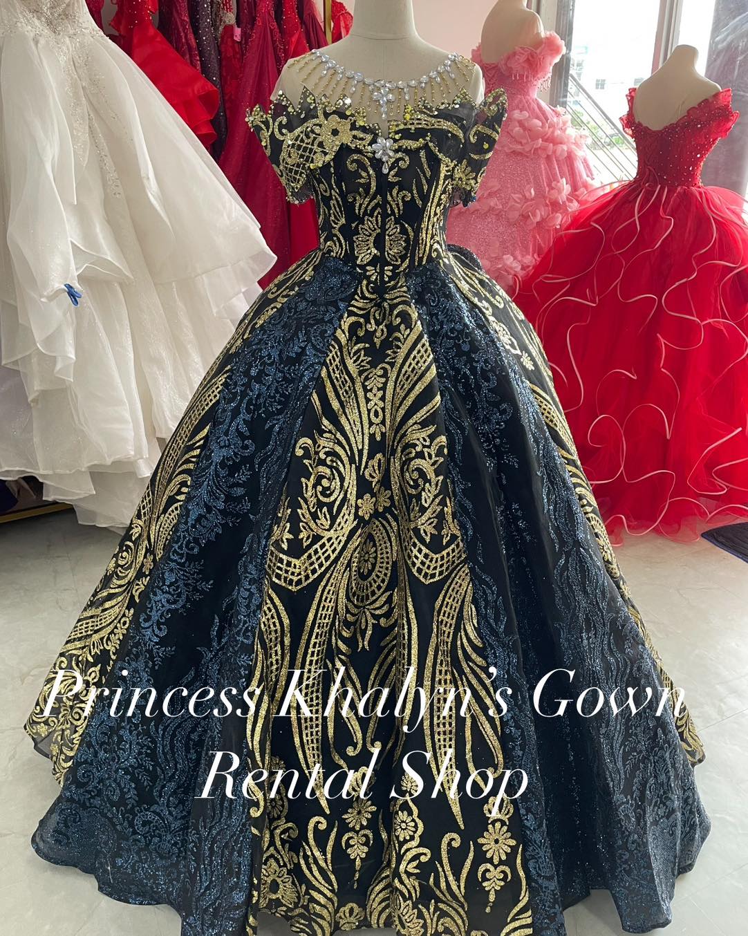 A luxurious black ball gown with intricate gold embroidery and a voluminous skirt. The gown features an off-shoulder design with ornate detailing and a pearl-embellished neckline, creating a regal and dramatic appearance.