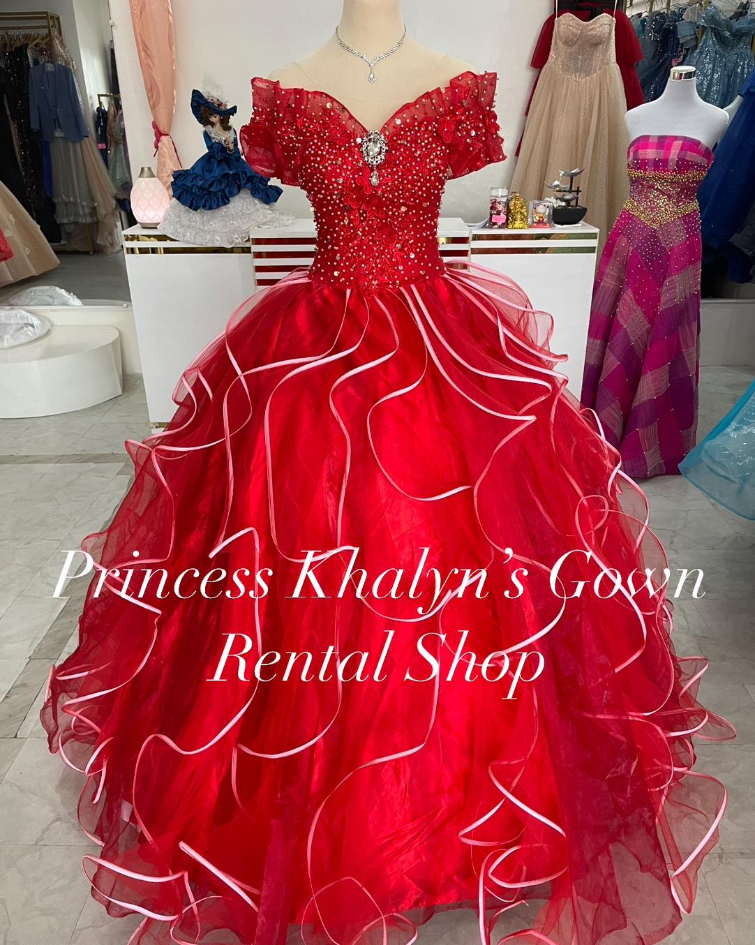 A vibrant red off-shoulder gown featuring a fitted bodice adorned with sparkling beadwork and a large central brooch. The voluminous skirt is accented with cascading ruffles and white trim, creating a dramatic and elegant silhouette.