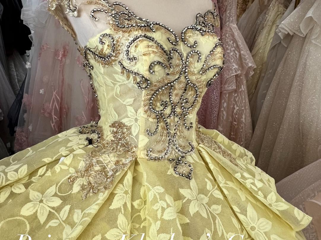 A luxurious gold gown featuring a fitted bodice adorned with intricate silver and gold beadwork and embroidery. The gown has an off-shoulder design with a voluminous skirt embellished with floral patterns, creating an elegant and sophisticated look.
