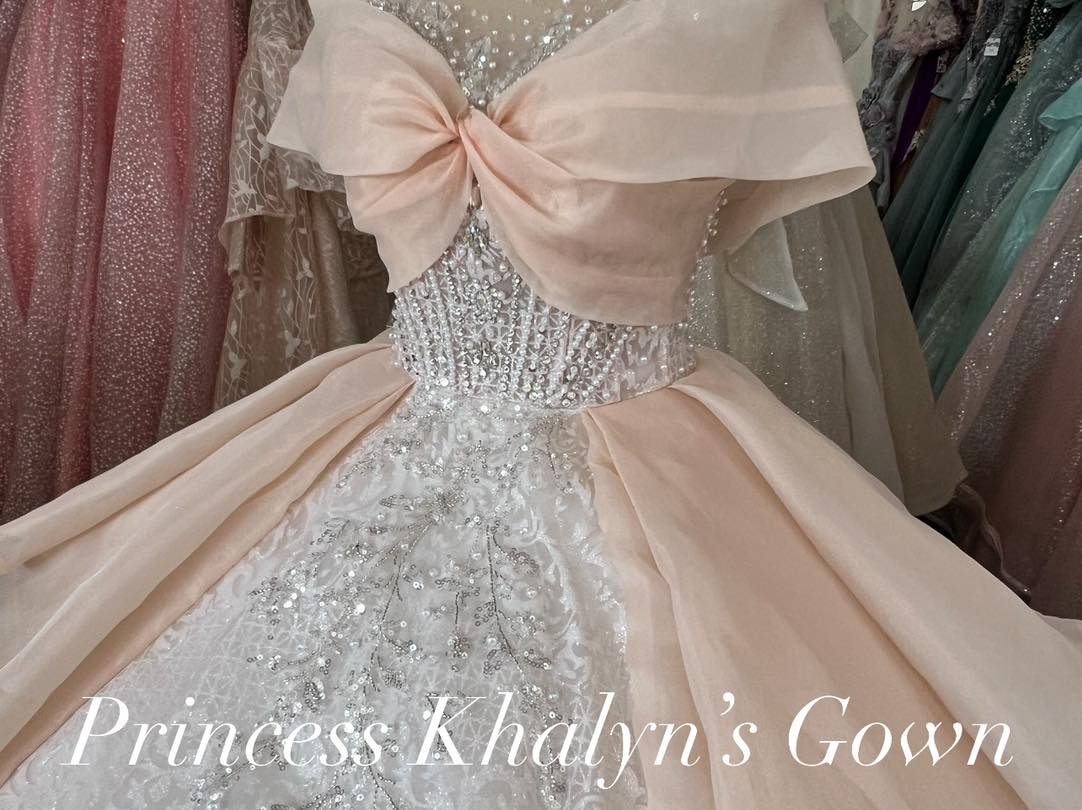A blush pink ball gown with a sheer embellished bodice, featuring a large bow at the neckline and a voluminous skirt adorned with intricate beadwork and lace detailing.
