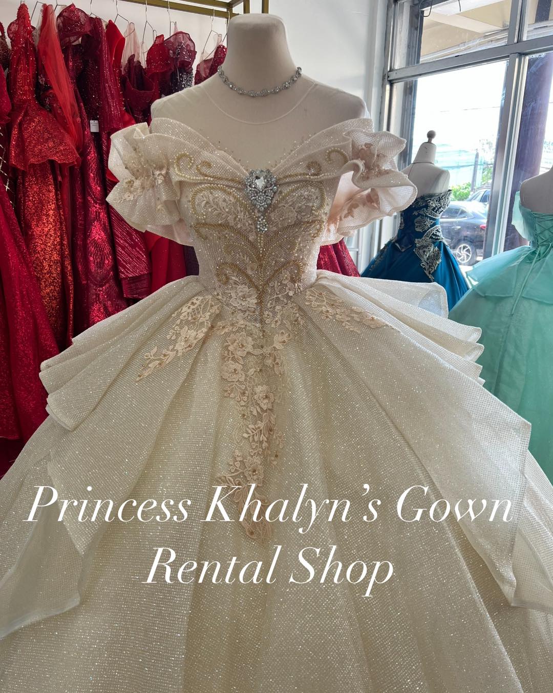 An elegant ivory off-shoulder ball gown with intricate gold embroidery and floral appliqué on the bodice. The gown features a voluminous shimmering skirt with layered detailing and delicate ruffle sleeves, adorned with a sparkling gemstone centerpiece.