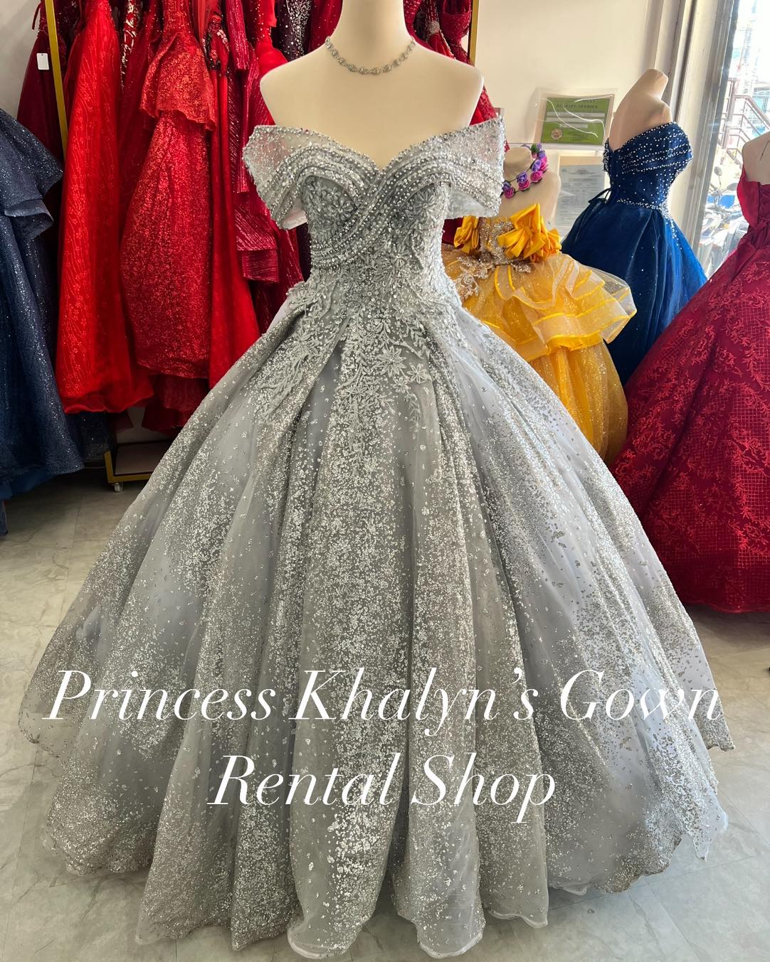 A stunning silver off-shoulder ball gown with intricate beadwork and embroidery on the bodice. The gown features a voluminous shimmering tulle skirt, creating a sparkling and elegant appearance.