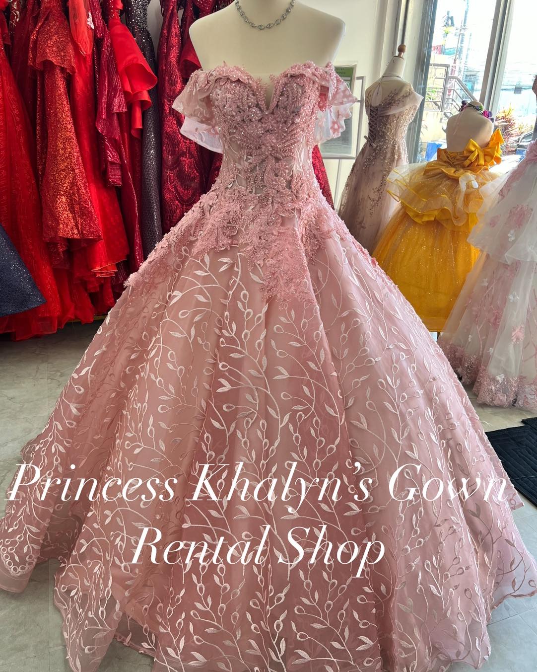 A pink off-shoulder ball gown with intricate floral lace embroidery on the bodice and a voluminous skirt adorned with delicate vine patterns. The gown features a sweetheart neckline and sheer ruffled sleeves, creating a romantic and elegant appearance.