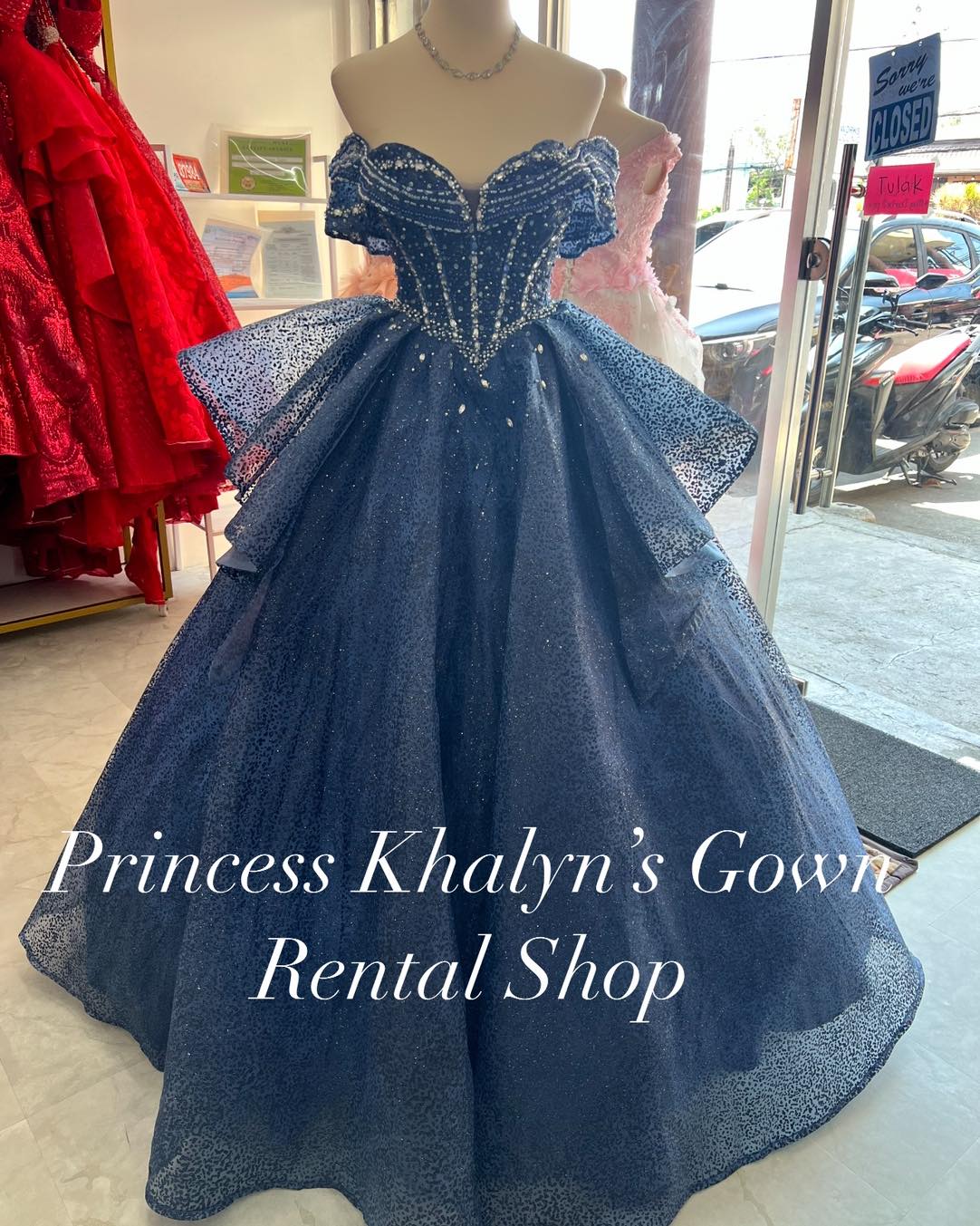 A navy blue off-shoulder ball gown with a shimmering bodice adorned with intricate beadwork and a voluminous glittery tulle skirt. The gown features a structured sweetheart neckline and elegant detailing, suitable for formal occasions.