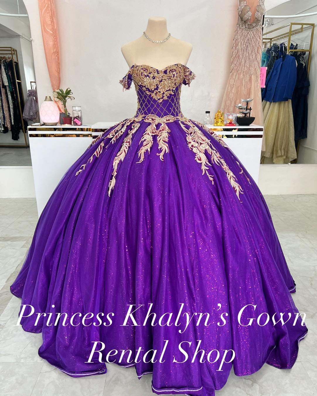 A luxurious purple ball gown with off-shoulder sleeves and intricate gold embroidery on the bodice. The gown features a voluminous skirt with shimmering fabric, creating an elegant and regal appearance.