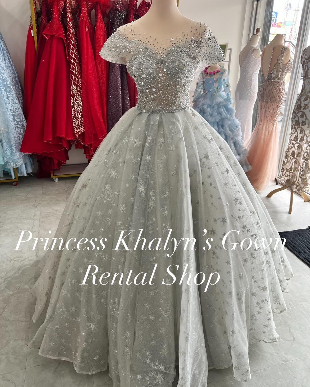 A stunning silver ball gown with an off-shoulder design, featuring intricate sequin and bead embellishments on the bodice and a voluminous tulle skirt adorned with star patterns. The gown exudes elegance and is suitable for formal occasions.