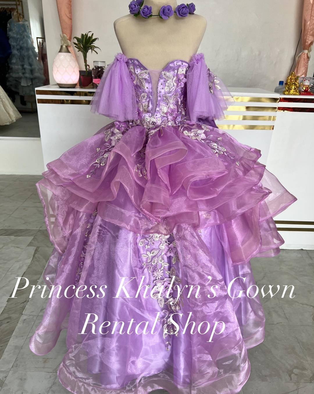 A stunning lavender purple ball gown featuring a sweetheart neckline with intricate floral embroidery. The gown has off-shoulder tulle sleeves and a voluminous layered skirt with ruffled detailing, creating a whimsical and enchanting appearance.