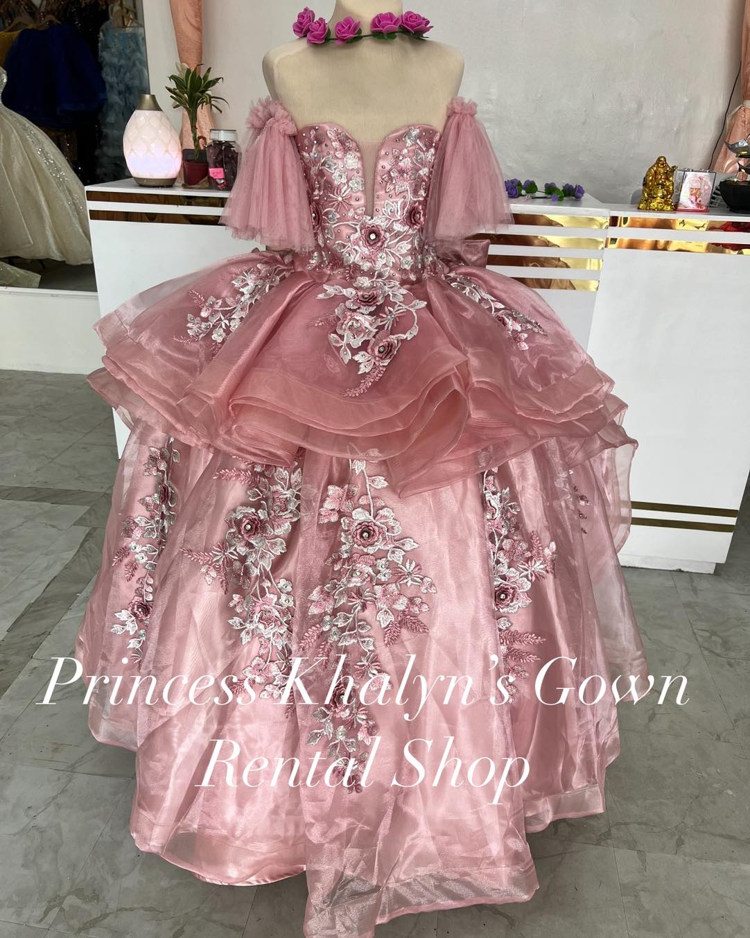 A stunning pink ball gown with off-shoulder puffed sleeves and intricate floral embroidery. The gown features a layered tulle skirt and a fitted bodice adorned with delicate lace and beadwork, creating a romantic and elegant silhouette.