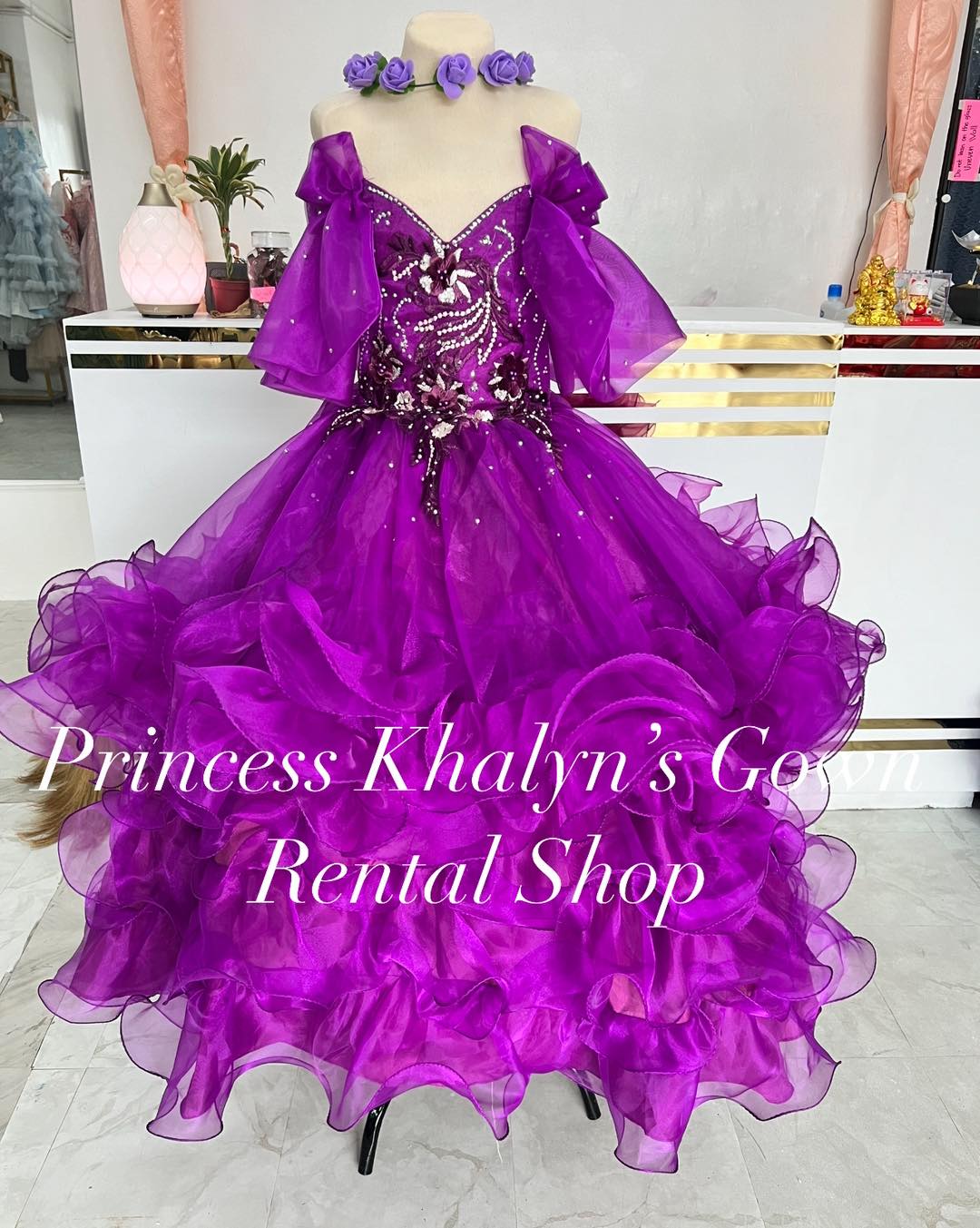 A vibrant purple ball gown with off-shoulder ruffled sleeves, intricate beadwork, and floral appliqué on the bodice. The skirt is voluminous with layers of ruffled tulle, creating a dramatic and enchanting silhouette.
