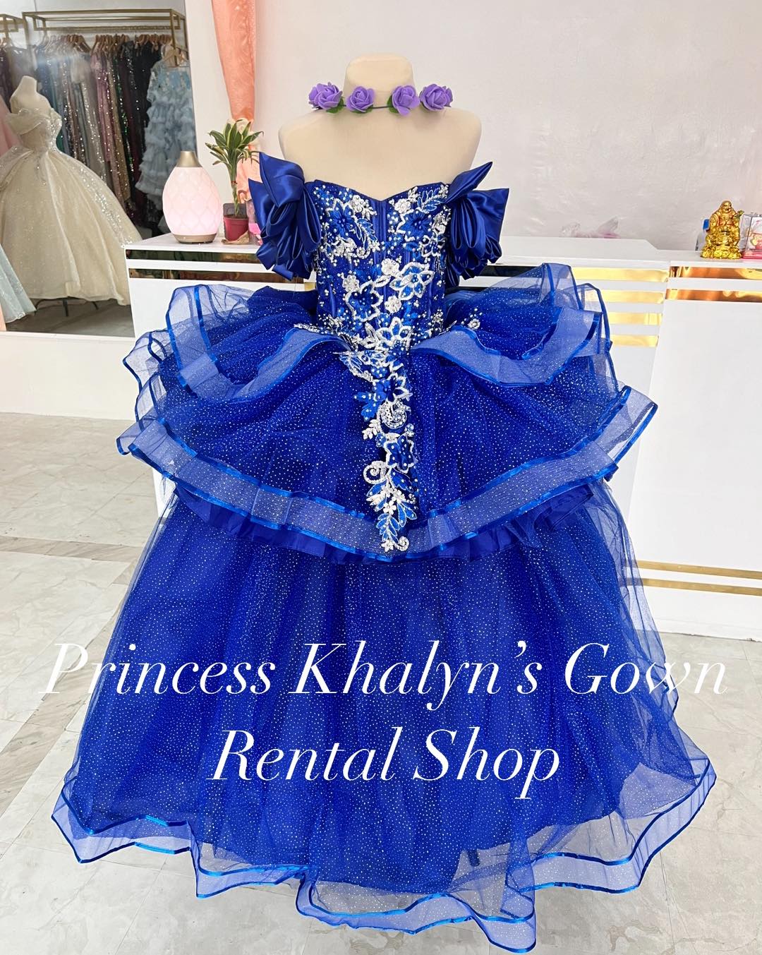 A vibrant royal blue ball gown with off-shoulder puffed sleeves, intricate silver floral embroidery, and a voluminous layered tulle skirt adorned with shimmering embellishments.