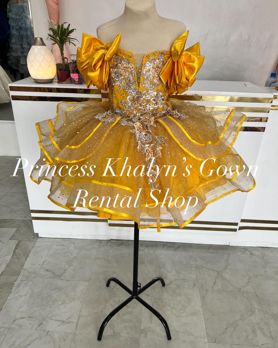 A vibrant gold gown with off-shoulder design, featuring large satin bows on the shoulders and intricate silver floral embroidery on the bodice. The skirt is layered with shimmering tulle, creating a sparkling and elegant appearance.