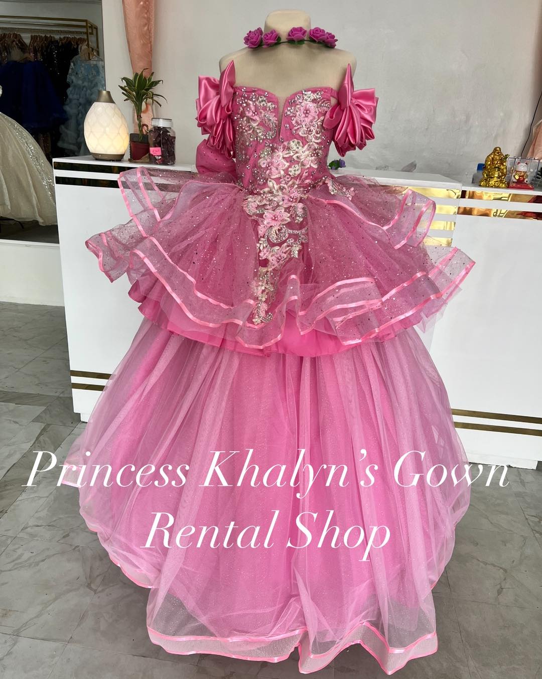 A vibrant pink ball gown with off-shoulder design, featuring layered ruffles and intricate floral embroidery on the bodice. The gown is adorned with sparkling embellishments and has a voluminous tulle skirt, creating a whimsical and elegant appearance.