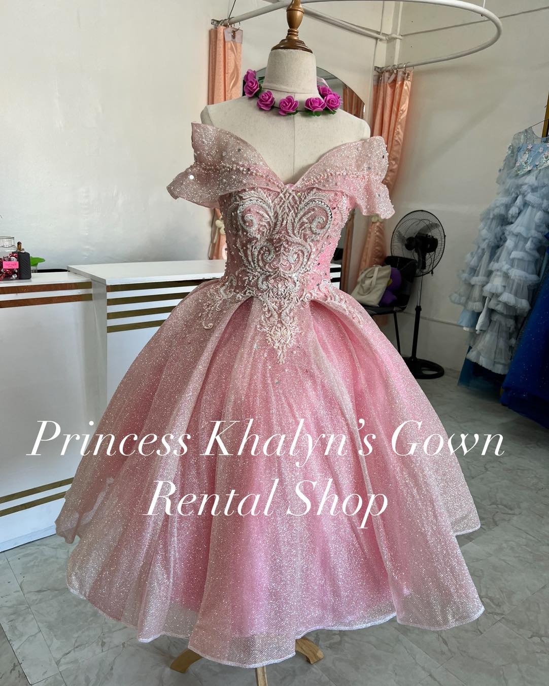 A shimmering pink off-shoulder ball gown with intricate silver embroidery on the bodice and a voluminous sparkling tulle skirt. The gown features delicate ruffle detailing on the sleeves, creating an enchanting and elegant look.