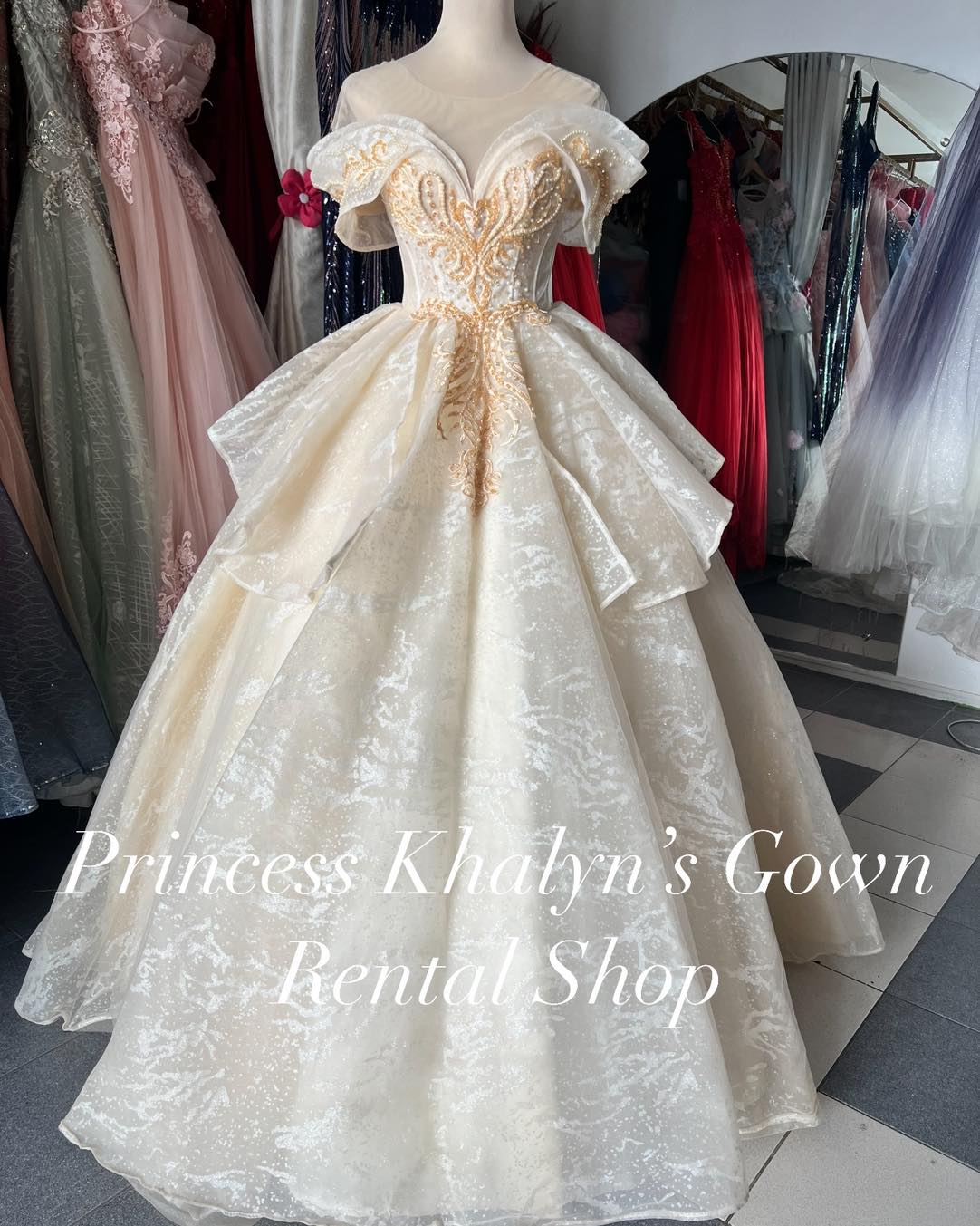 An elegant ivory ball gown with an off-shoulder design, featuring intricate gold embroidery on the bodice and a voluminous shimmering skirt with layered detailing. Perfect for weddings, formal events, and debuts.