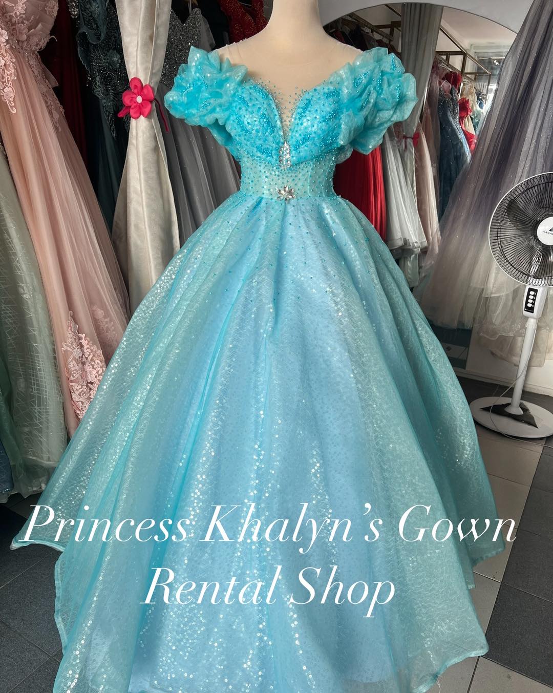 A dazzling aqua blue ball gown with off-shoulder puffed sleeves, intricate beadwork on the bodice, and a shimmering full skirt. The gown features a delicate floral embellishment at the waist, creating a whimsical and enchanting look.