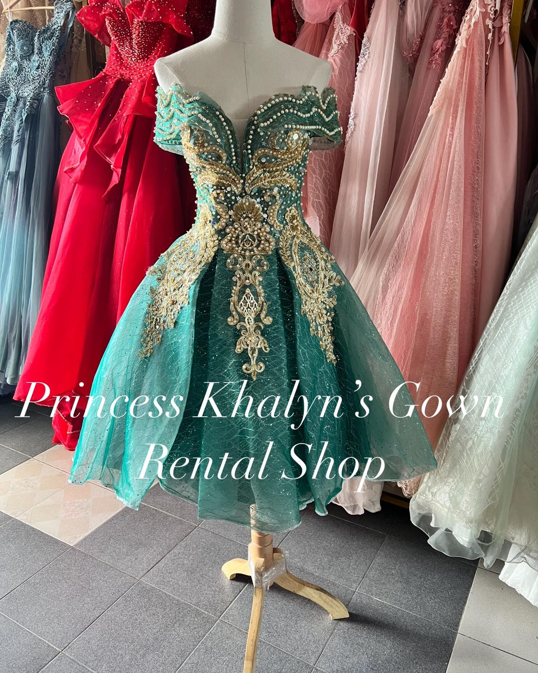 A stunning emerald green off-shoulder gown with intricate gold embroidery and beadwork on the bodice. The gown features a sweetheart neckline and a short, voluminous skirt with a shimmering overlay, perfect for formal occasions.