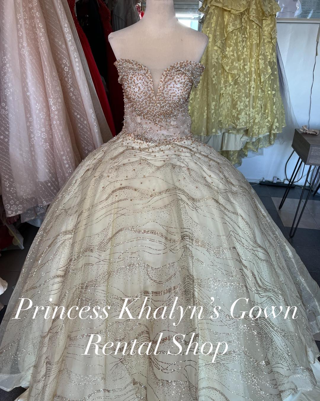 A luxurious champagne gold ball gown with an off-shoulder design. The bodice is adorned with intricate beadwork and sequins, creating a shimmering effect. The voluminous skirt features delicate wave-like patterns of glitter, adding to the gown's opulent appearance.
