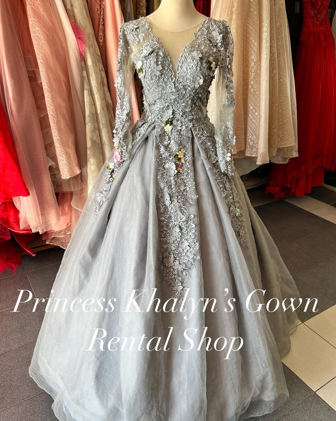 A silver ball gown with a deep V-neckline and long sheer sleeves. The gown features intricate floral lace embroidery on the bodice and sleeves, with colorful floral accents. The voluminous skirt adds a dramatic flair, making it suitable for weddings, formal events, and parties.