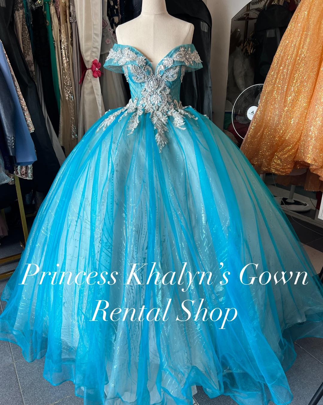 A dazzling aqua blue off-shoulder ball gown with intricate silver embroidery and beadwork on the bodice. The gown features a voluminous shimmering tulle skirt, creating an ethereal and elegant appearance.