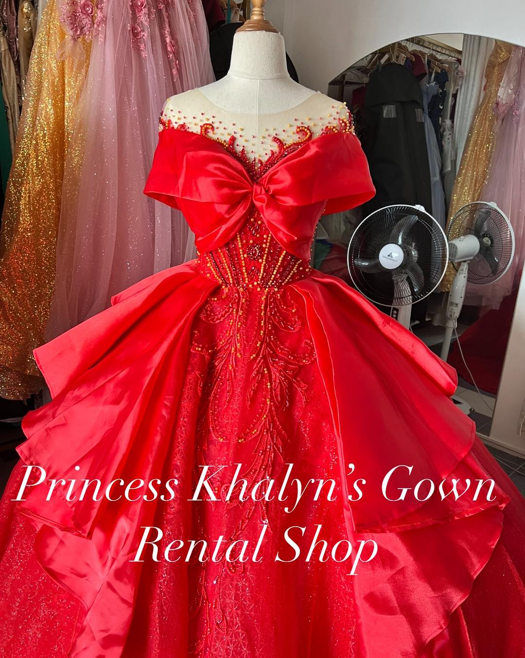 A vibrant red ball gown with an off-shoulder design, featuring a large decorative bow on the bodice and intricate beadwork. The gown has a voluminous skirt with shimmering details, creating a dramatic and elegant appearance.