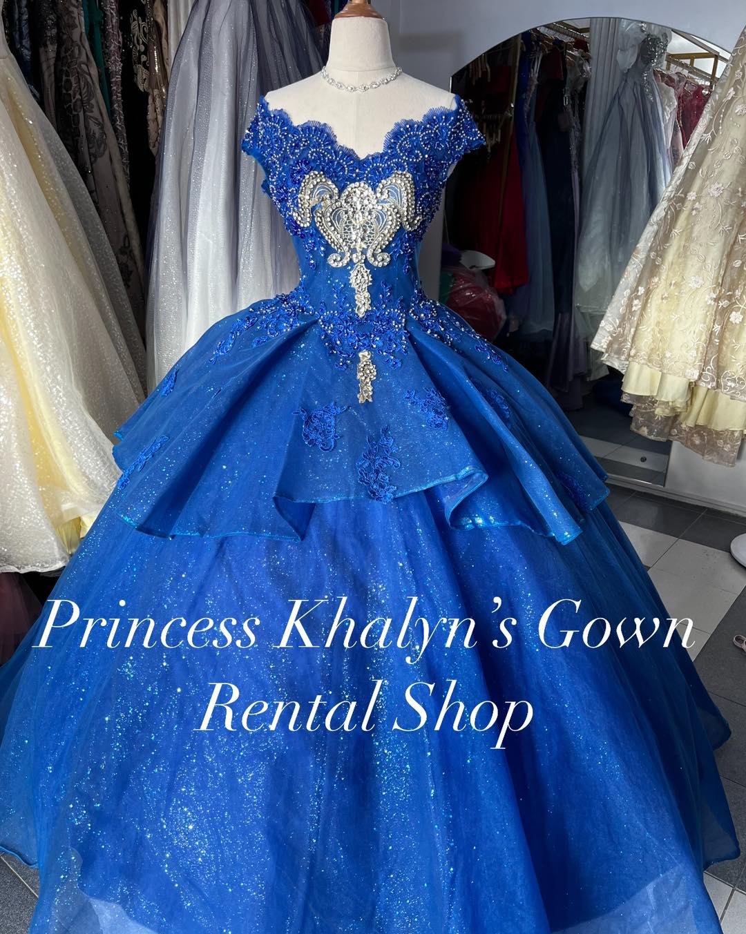 A dazzling royal blue ball gown featuring an off-shoulder design with intricate silver embroidery and beadwork on the bodice. The voluminous skirt is adorned with shimmering fabric, creating a majestic and elegant appearance.