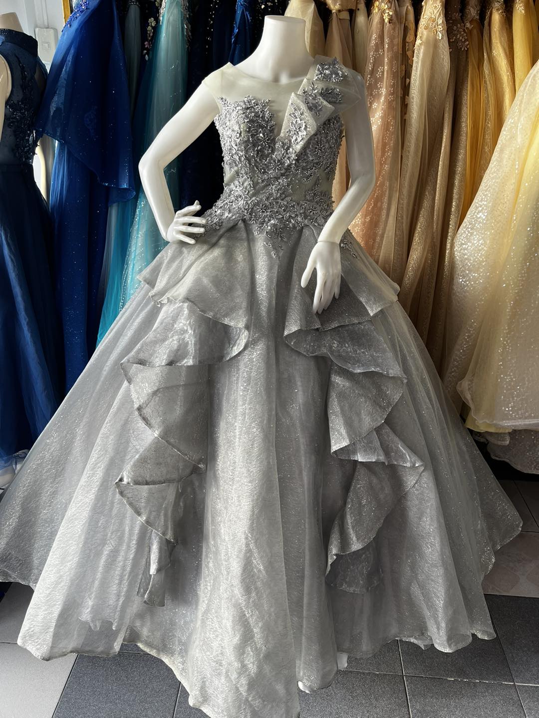 A stunning silver gown with a sheer neckline and intricate floral embroidery on the bodice. The gown features cascading ruffles on the voluminous skirt, creating an elegant and sophisticated look.